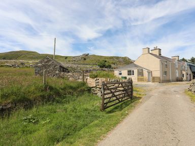 A driveway with a wooden gate leading to a house and a small stone building in rural landscape at Cwm Ceiliog Annex in Llanaelhaearn near Trefor