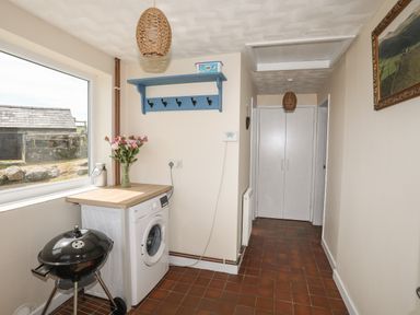 A hallway with a washing machine and countertop a black charcoal grill a blue wall-mounted shelf with hooks and a window with a view of an outdoor stone wall at Cwm Ceiliog Annex in Llanaelhaearn near Trefor