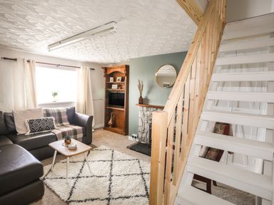 A living room with a brown leather sofa a small wooden table a white rug a wooden cabinet with a TV and a staircase with wooden railing at Cwm Ceiliog Annex in Llanaelhaearn near Trefor