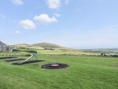 A playground with a slide swings and a roundabout on a grassy field with hills in the background at Cwm Ceiliog Annex in Llanaelhaearn near Trefor