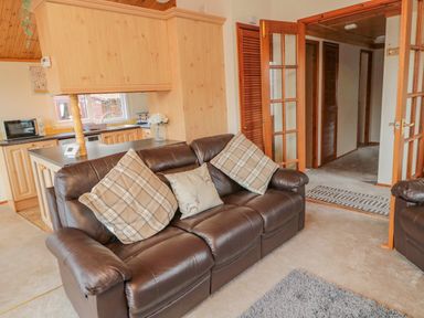 A living area with a brown leather sofa and plaid cushions next to a kitchen with wood cabinets at 4 The Oaks in Swarland