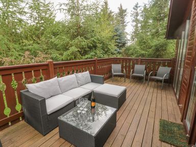 An outdoor wooden deck with a sofa set coffee table with glasses and a bottle and three chairs surrounded by trees at 4 The Oaks in Swarland