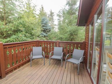 A wooden deck with three gray chairs and a wooden railing surrounded by trees at 4 The Oaks in Swarland