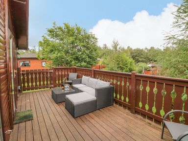 An outdoor wooden deck with a wicker sofa set and trees in the background at 4 The Oaks in Swarland