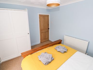 A bedroom with a wooden bed frame and yellow bedding and folded grey towels on the bed and a white wardrobe and wooden door at Ty Cwtch in Tenby