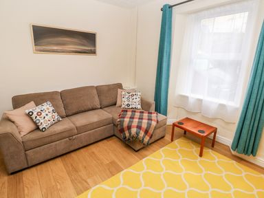 A living room with a brown sectional sofa with pillows and a blanket a wooden small table yellow patterned rug and blue curtains at Ty Cwtch in Tenby
