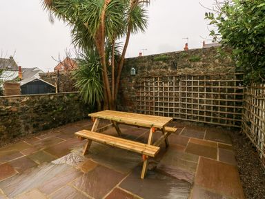 An outdoor patio area with a wooden picnic table and benches surrounded by stone walls and plants at Ty Cwtch in Tenby