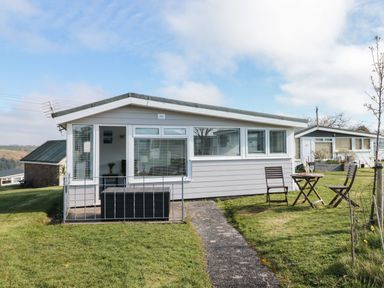 A single-story house with white siding and multiple windows with a small patio and outdoor table with two chairs on grass at Wagtails in Norton near Dartmouth