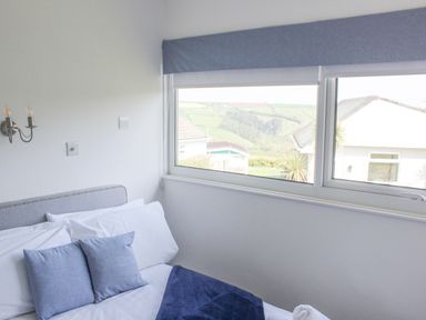 A bedroom with a bed with white and blue pillows and a folded towel near a window showing houses and hills at Wagtails in Norton near Dartmouth