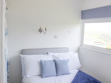 A bedroom with a bed with white and blue pillows a window showing a view outside at Wagtails in Norton near Dartmouth