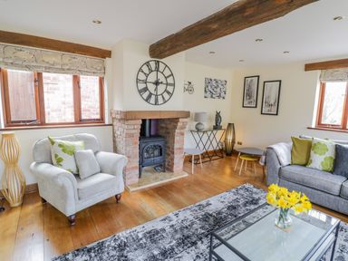 A living room with armchair and sofa near a brick fireplace with clock above and flowers on glass table at The Stables in Belbroughton