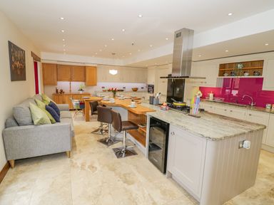 An open kitchen with a large island, bar stools, a sofa with cushions, wooden cabinets, and a pink backsplash at The Stables in Belbroughton