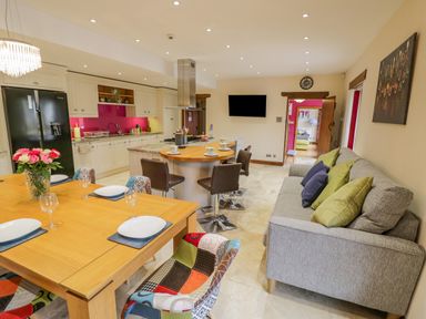 An open kitchen and dining area with a wooden table, chairs, a grey sofa with cushions, kitchen island with stools and a wall-mounted tv at The Stables in Belbroughton