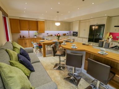 A kitchen and dining area with wooden cabinets a black refrigerator a wooden dining table set with plates and cups and a gray sofa with green and blue cushions at The Stables in Belbroughton