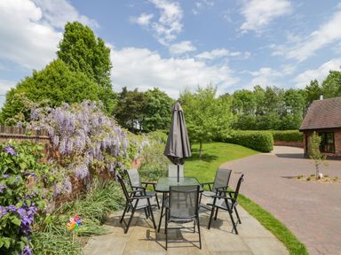 An outdoor patio with a glass table and six chairs with a closed umbrella near a brick wall with flowers and a garden at The Stables in Belbroughton