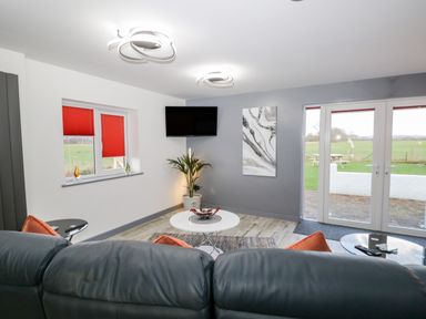 A sitting area with a black leather sofa and orange cushions a white round coffee table a plant a wall-mounted tv and large glass doors at The Annex in Ulverston