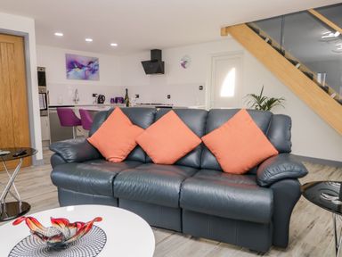 A living room with a black leather sofa and orange cushions near a staircase and kitchen at The Annex in Ulverston