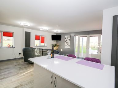 A kitchen island with two purple chairs and a bottle with glasses facing a living room with a black sofa and a wall-mounted tv with glass doors leading outside at The Annex in Ulverston