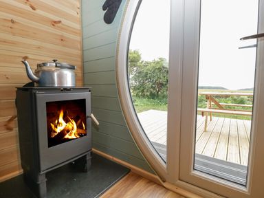 A wood burning stove with a kettle on top next to a round window and glass door with a view of a deck and picnic table at Dew Pond in Blagdon