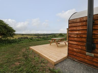 An outdoor wooden deck with a picnic table next to a wooden structure with a stove pipe and fields in the background at Dew Pond in Blagdon