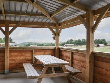 A wooden picnic table and benches under a covered wooden shelter overlooking a rural landscape at Dew Pond in Blagdon