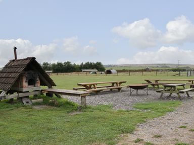 An outdoor seating area with wooden picnic tables a fire pit and a brick pizza oven in a field at Dew Pond in Blagdon