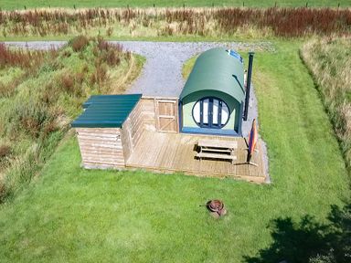 A small green curved cabin with a wooden deck picnic table and umbrella on grass at Dew Pond in Blagdon