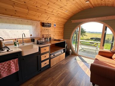 A small kitchen and living area with wooden walls and ceiling a sofa and round glass door opening to a patio at Dew Pond in Blagdon