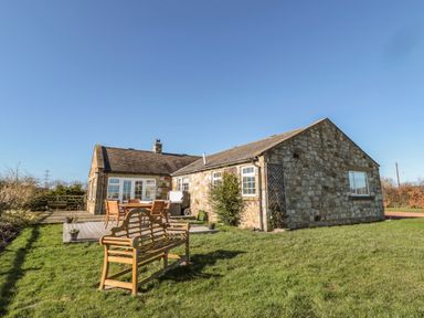 A stone house with a wooden bench and outdoor dining set on a lawn at Thistleyhaugh Cottage in Longhorsley near Longframlington