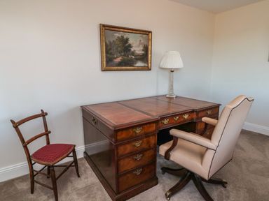 An office room with a large wooden desk a beige office chair wooden chair with red cushion lamp and landscape painting on the wall at Thistleyhaugh Cottage in Longhorsley near Longframlington