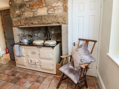 A kitchen with stone wall fireplace cooker kettle wooden chair with cushions at Thistleyhaugh Cottage in Longhorsley near Longframlington