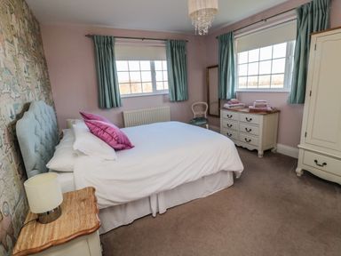 A bedroom with a bed with white linens and pink pillows bedside tables a chest of drawers a chair and windows with green curtains at Thistleyhaugh Cottage Longhorsley near Longframlington