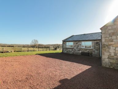 A stone cottage with two windows facing a gravel courtyard and a fenced field in the background at Thistleyhaugh Cottage Longhorsley near Longframlington