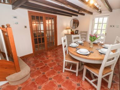 A dining room with a round wooden table set for six and white chairs on a tiled floor at Trevedran Cottage in St Buryan