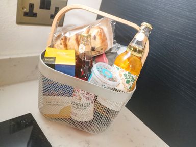A small basket with bottles drinks, a box of clotted cream shortbread, and packaged pastries on a kitchen counter at Trevedran Cottage in St Buryan