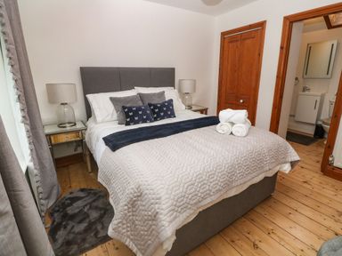 A bedroom with a bed featuring pillows and a blanket bedside tables with lamps wooden floors and a door leading to a bathroom at Trevedran Cottage in St Buryan