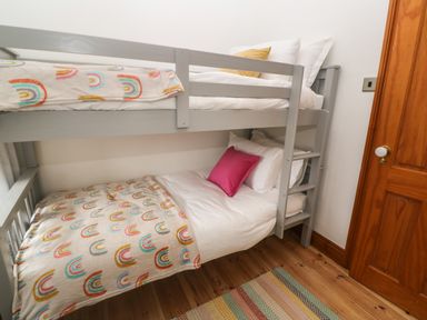 A bedroom with a gray bunk bed with white bedding and colorful blankets next to a wooden door on a wooden floor at Trevedran Cottage in St Buryan
