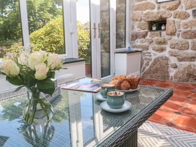 A glass table with a vase of flowers two cups of coffee and pastries in a room with a stone wall at Trevedran Cottage in St Buryan