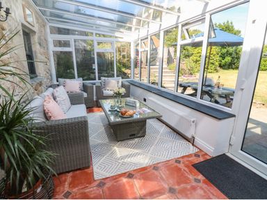 A conservatory with wicker furniture coffee table and large windows overlooking a garden at Trevedran Cottage in St Buryan