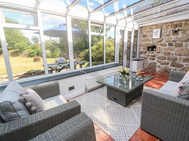 A sunroom with wicker sofas a glass coffee table and a rug overlooking a garden at Trevedran Cottage in St Buryan