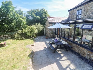 A stone house with large windows and a patio featuring a picnic table with an umbrella and a garden with bushes at Trevedran Cottage in St Buryan