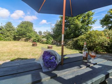 A picnic table with a bouquet of purple flowers a bottle and glass of wine under an umbrella in a garden at Trevedran Cottage in St Buryan
