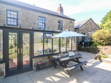 A stone house with large windows and a patio table with umbrella on a tiled patio at Trevedran Cottage in St Buryan
