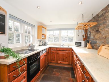 A kitchen with wooden cabinets countertops a built in oven stovetop microwave sink windows and stone wall at Bay Tree Cottage Ambleside