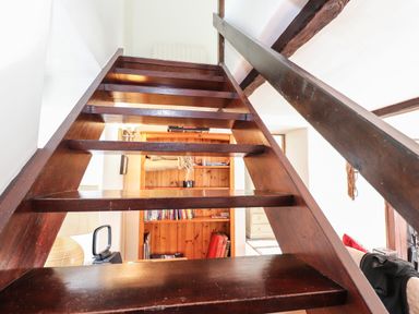 A wooden staircase with open risers overlooking a room with a wooden bookshelf and white walls at Bay Tree Cottage in Ambleside