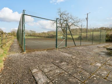 An outdoor tennis court surrounded by chain link fencing next to a gravel pathway and a picnic bench at Orchard Lodge in Hockworthy near Sampford Peverell Devon