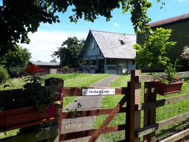 A wooden gate with flowers on it leading to a house with a triangular roof at Orchard Lodge in Hockworthy near Sampford Peverell Devon