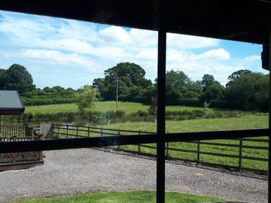 A countryside view through a window showing a fenced grassy field with trees and a small pond at Orchard Lodge in Hockworthy near Sampford Peverell Devon