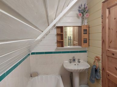 A bathroom with a sink and mirror under a sloped ceiling with a wooden door and flowers on a shelf at Orchard Lodge in Hockworthy near Sampford Peverell Devon