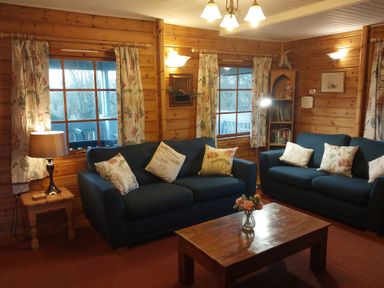 A living room with two blue sofas and floral cushions a wooden coffee table with flowers a wooden side table with a lamp and floral curtains at Orchard Lodge in Hockworthy near Sampford Peverell Devon